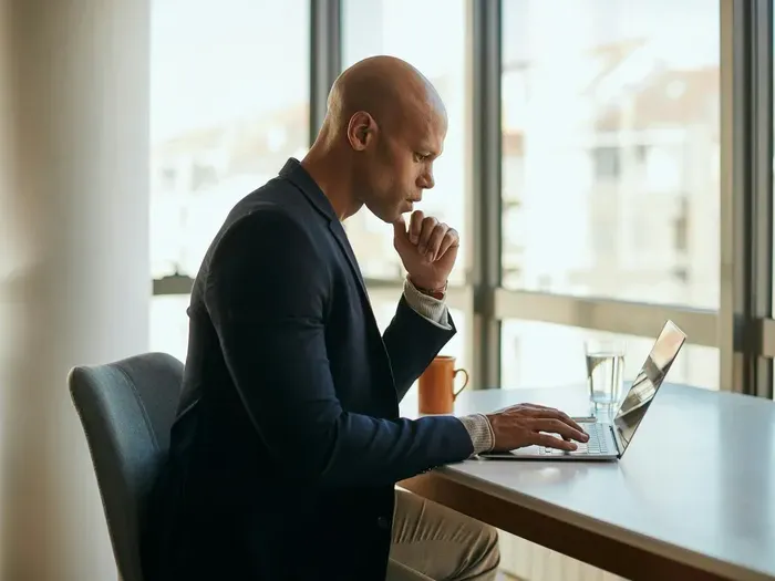 Focused businessman in formal suit working on a laptop at a bright modern office desk with glass of water nearby